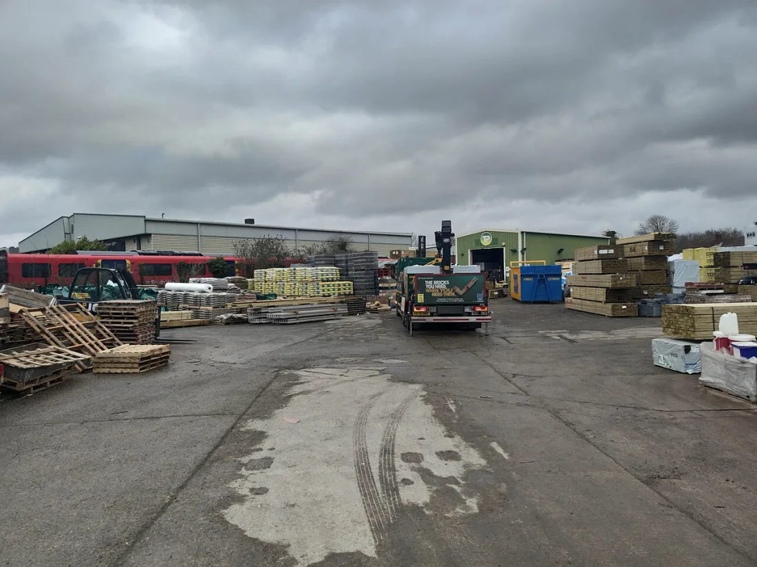 A UK builders merchant yard stacked with timber, bagged materials and aggregates, with a delivery lorry and forklift
